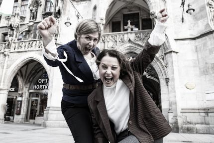 Zwei menschen in vage historisierenden Anzügen recken die Fäuste vor dem Münchner Rathaus, alles in Sepia